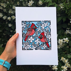 May include: A rectangular art print on white paper featuring a linocut-style illustration of two red cardinals perched among white flowers and black leaves against a light blue background with swirling patterns and a black border frame.