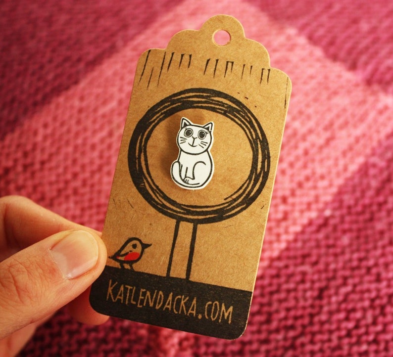 May include: A brown paper tag with a white cat illustration inside a circle. The tag has the text "KATLENDACKA.COM" printed on the bottom. A small red bird illustration is on the left side of the tag.