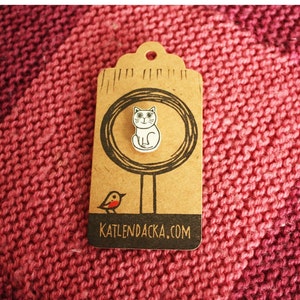May include: A white cat pin with black eyes and a black nose, sitting on a brown tag with a black circle and a black bird. The tag has the text "KATLENDACKA.COM" printed on it.