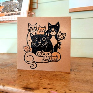 May include: A square greeting card with a brown background features a black and orange cat illustration. The illustration depicts a group of cats of various sizes, with some cats overlapping each other. The cats have simple, stylized features.