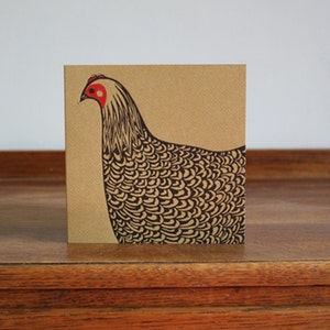 May include: A square greeting card featuring a stylized illustration of a hen. The hen is depicted in black and white with a red comb and wattle, set against a tan background. The card is displayed on a wooden surface.