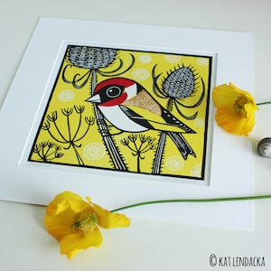 May include: A yellow and black linocut print of a bird with a red head and a yellow breast, surrounded by black and white stylized plants on a yellow background. The print is signed "Kat Lendaacka".