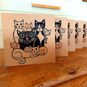 May include: A stack of square greeting cards with a black and orange cat illustration on a kraft paper background. The cat design features a large black cat surrounded by smaller cats in various poses. The cards are displayed on a wooden surface.