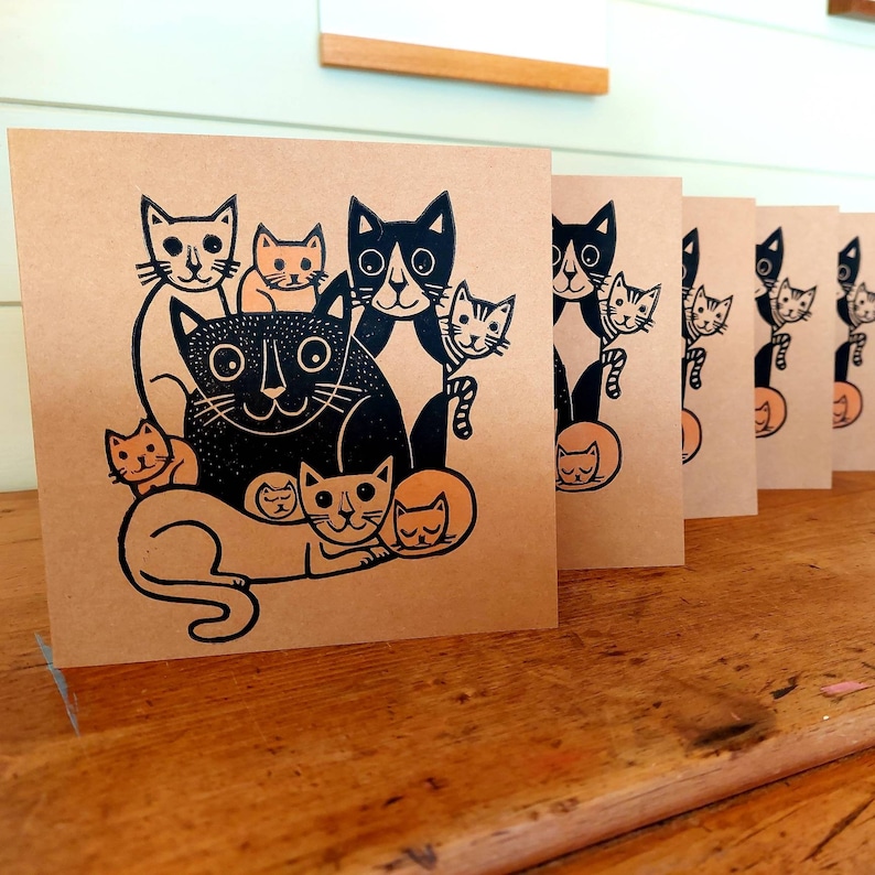 May include: A stack of square greeting cards with a black and orange cat illustration on a kraft paper background. The cat design features a large black cat surrounded by smaller cats in various poses. The cards are displayed on a wooden surface.
