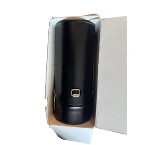 May include: A black cylindrical object with a small, rectangular gold-coloured detail, presented inside a partially open cardboard box. The object has a smooth, matte finish and a rounded base. The box is plain, with a neutral colour.
