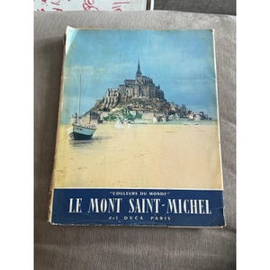 Op de afbeelding: Vintage reisboekcover met een schilderachtig uitzicht op Mont Saint-Michel, Frankrijk, onder een blauwe lucht. Een kleine boot drijft op de voorgrond. De titel "LE MONT SAINT-MICHEL" staat in witte tekst op een blauwe banner.