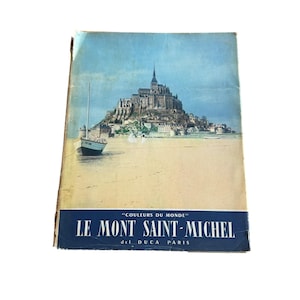 Op de afbeelding: Vintage reisbrochure met een schilderachtig uitzicht op Le Mont Saint-Michel, Frankrijk. De omslag toont een gedetailleerde illustratie van de abdij van het eiland en de omliggende wateren, met een kleine boot op de voorgrond. De titel is in het Frans.