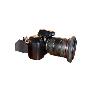 May include: Black Nikon DSLR camera with a large lens and a black strap. The lens has measurement markings. The camera is positioned on a light brown surface.