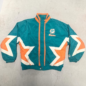 Puede incluir: Chaqueta vintage de color turquesa y naranja con detalles de estrellas blancas en las mangas. Cuenta con cierre de botones, cuello alzado y el logotipo de los Dolphins bordado en el pecho. Una chaqueta deportiva retro.