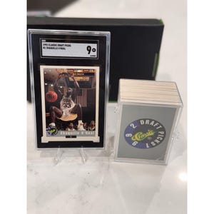 Puede incluir: Una tarjeta coleccionable de 1992 Classic Draft Picks con Shaquille O'Neal, calificada con un 9. La tarjeta está en un soporte negro con un soporte transparente. Una pila de tarjetas similares está en una funda de plástico transparente.