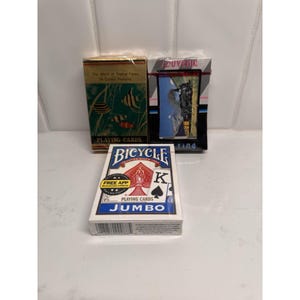 Puede incluir: Se muestran tres barajas de cartas. Una es una baraja Bicycle Jumbo con un diseño rojo y blanco. Las otras dos barajas tienen gráficos y texto coloridos, incluyendo "The World of Tropical Fishes" y una imagen de recuerdo.