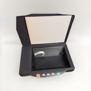 Puede incluir: Un escáner de superficie plana negro con la tapa abierta, que revela un interior beige y una superficie de escaneo de vidrio. El escáner tiene varios botones en la parte frontal y está sobre un fondo blanco.