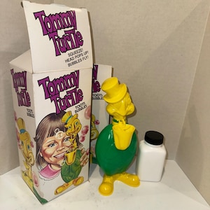 Peut inclure: Un ensemble de jouets à bulles Tommy Turtle vintage. L'ensemble comprend un souffleur de bulles en forme de tortue verte avec des accents jaunes, une bouteille blanche et une boîte avec le texte "Tommy Turtle" et une illustration d'une fille et d'une tortue.