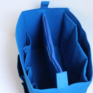 May include: A bright blue fabric organizer insert with a structured rectangular shape. The interior features a central zippered compartment, multiple vertical side pockets, and open storage sections designed to fit inside a larger bag.