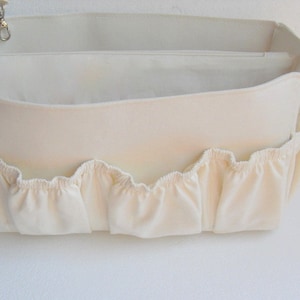 May include: A white canvas bag organiser with multiple compartments and a ruffled edge. The bag has a zippered top and a small loop for attaching a key chain.