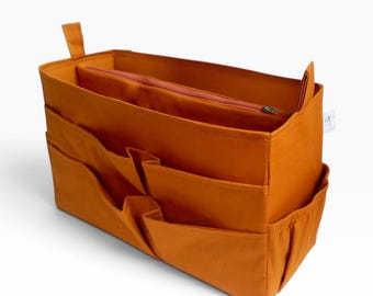 May include: A rectangular burnt orange felt bag organizer featuring multiple exterior slip pockets, a central zippered compartment, and two small fabric tabs on the top corners. A small white rectangular tag is attached to the side.