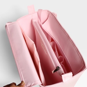 Peut inclure: Un organisateur de sac en tissu rose clair avec une ouverture sur le dessus, comprenant plusieurs compartiments intérieurs, des poches latérales et une section centrale zippée. La structure est rectangulaire avec des bords renforcés.