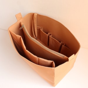 Op de afbeelding: Een beige stoffen tasorganizer met meerdere vakken en een ritssluiting. De organizer is ontworpen om in een grotere tas te passen en helpt om spullen georganiseerd te houden.