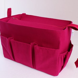 Peut inclure: Un organisateur de sac fourre-tout magenta avec plusieurs poches extérieures et un compartiment supérieur zippé. Le sac a une poignée sur le côté et une petite boucle sur le dessus. L'organisateur est conçu pour être placé dans un sac plus grand.