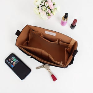 Op de afbeelding: Zwarte handtassen organizer met meerdere compartimenten en een bruin interieur. De organizer is open en toont vakken en een gedeelte met ritssluiting. Een smartphone, sleutels en cosmetica zijn in de buurt geplaatst. Een klein boeket bloemen staat op de achtergrond.