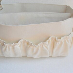 May include: A white canvas bag organiser with multiple compartments and a ruffled edge. The bag has a zippered top and a small loop for attaching a key chain.