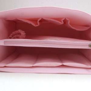 Op de afbeelding: Een lichtroze draagtas met meerdere compartimenten en een vak met ritssluiting. De binnenkant heeft verschillende zakken en een scrunchie. De tas is ontworpen voor organisatie en opslag.