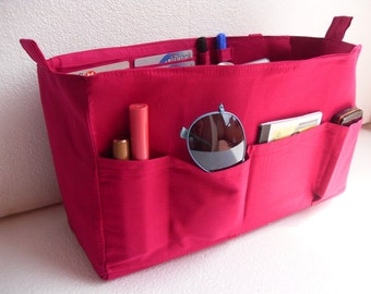 May include: A bright pink fabric organizer with multiple pockets and handles. The organizer is filled with various items, including sunglasses, lipstick, pens, credit cards, and a small notebook. The pockets are designed to hold small items for easy access.