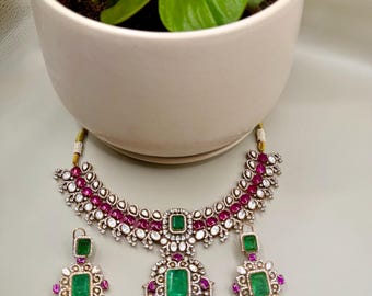 May include: A silver-toned jewelry set featuring a necklace and matching earrings. The necklace and earrings are adorned with emerald-green and ruby-red gemstones, and clear crystals. The set is displayed near a white ceramic pot with a green plant.