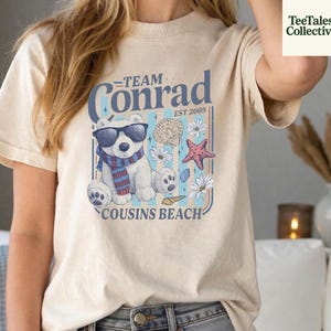 May include: Beige t-shirt with a graphic featuring a polar bear wearing sunglasses and a scarf, surrounded by seashells and flowers. Text reads "Team Conrad, Cousins Beach, Est 2009."