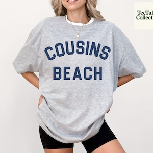 Puede incluir: Una sudadera gris claro con las palabras "COUSINS BEACH" en letras azul marino. La sudadera tiene cuello redondo y mangas cortas. La imagen también incluye pantalones cortos negros.