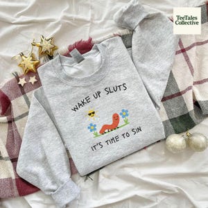 Peut inclure: Un sweat-shirt gris clair avec le texte "WAKE UP SLUTS IT'S TIME TO SIN" et un dessin animé d'un soleil souriant, d'un ver et de fleurs. Le sweat-shirt est plié sur une surface blanche avec des étoiles décoratives et une couverture à carreaux.