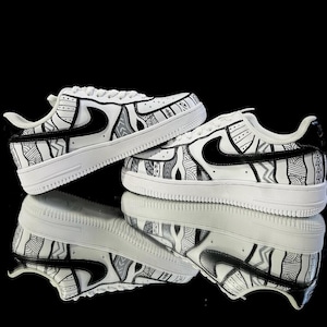 May include: Pair of white sneakers with black and gray tribal patterns. The shoes feature a black swoosh logo and are set against a reflective black surface. The design includes geometric and abstract shapes.