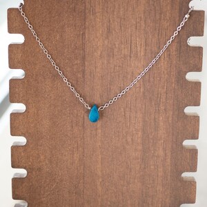 May include: A delicate silver necklace featuring a small, teardrop-shaped turquoise pendant. The necklace is displayed on a wooden stand with a natural finish, showcasing the jewelry's simple elegance. The chain is fine and the pendant is a vibrant blue.