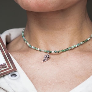 Può includere: Una collana di perline verdi e bianche con un ciondolo a forma di foglia d'argento. La collana è indossata. Si tiene in mano una carta con un'immagine verde e marrone.