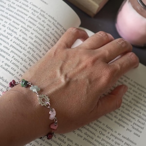 May include: A gemstone bracelet with pink, green, and red stones is shown on a wrist. The bracelet is made of silver-coloured metal. The bracelet is next to an open book.