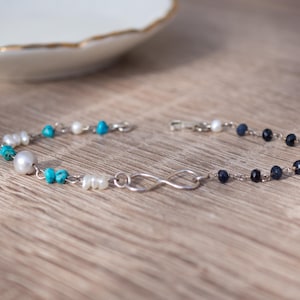 May include: A delicate bracelet featuring an infinity symbol and a mix of beads. The bracelet incorporates white pearls, turquoise, and dark blue beads, strung on a silver chain. The bracelet is displayed on a wooden surface.