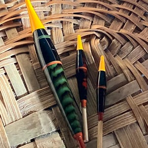 Könnte beinhalten: Drei Angelposen mit gelben Spitzen und verschiedenen Farben. Die größte Pose hat ein grün-schwarzes Muster, während die kleineren rote und schwarze Akzente aufweisen. Die Posen liegen auf einem geflochtenen Korb.