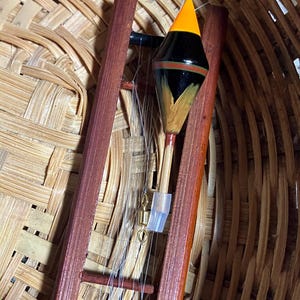 Puede incluir: Un flotador de pesca con un diseño negro, naranja y amarillo, sujeto a un marco de madera. El flotador tiene forma de lágrima y está suspendido por un hilo de pescar. El marco es de madera oscura y está sobre un fondo tejido.