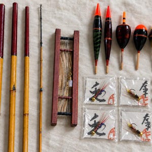 May include: A collection of fishing gear including three bamboo rod sections, a thin rod tip, a wooden line winder with line, five colorful fishing floats, four small plastic packets containing fishing flies with Japanese text, and various lead sinkers.