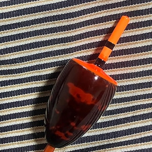 May include: A vintage fishing bobber with a dark brown, tortoiseshell-patterned body and an orange top. The bobber has a wooden stem and is accented with black stripes. The background is a striped fabric.