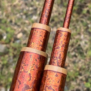Puede incluir: Primer plano de dos varillas de color cobre con un patrón texturizado y moteado en tonos naranja y marrón. Las varillas tienen forma cilíndrica con una banda marrón claro cerca de la base y la parte superior. El fondo está borroso.