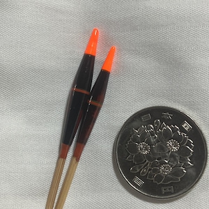 May include: Two black and orange fishing floats with a wooden base, next to a Japanese coin. The floats have a tapered shape with orange tips and red accents. The coin features a floral design.