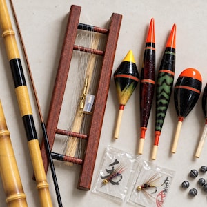 May include: A collection of fishing gear including two bamboo rod sections, a wooden line winder with clear fishing line, five colorful painted fishing floats, two small plastic bags containing feathered fishing hooks, and several small lead sinkers.