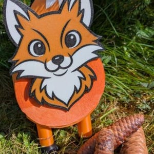 Könnte beinhalten: Eine dekorative orangefarbene Fuchsskulptur mit weißer und schwarzer Kontur, stehend auf orangefarbenen Beinen. Der Fuchs hat ein lächelndes Gesicht und steht neben drei braunen Tannenzapfen auf grünem Gras.