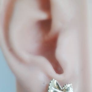 May include: Gold-toned earrings shaped like a bow tie pasta, also known as farfalle pasta. The earrings are shown in a person's ear.