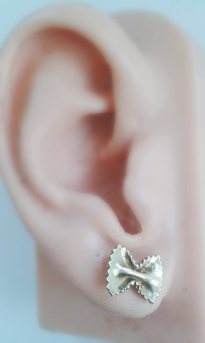 May include: Gold-toned earrings shaped like a bow tie pasta, also known as farfalle pasta. The earrings are shown in a person's ear.