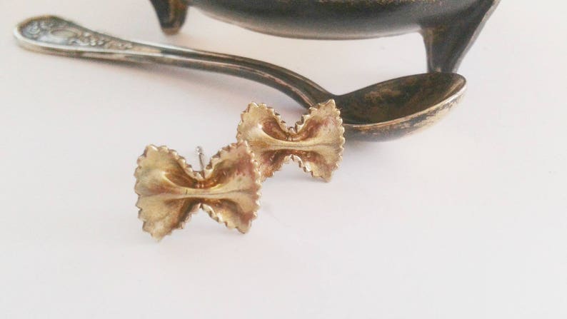 May include: A pair of gold-toned earrings shaped like pasta bows. The earrings are delicate and have a simple design.
