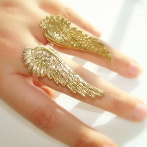 Könnte beinhalten: Ein goldfarbener Ring mit zwei Engelsflügel-Designs. Die Flügel sind detailliert und haben eine strukturierte Oberfläche.