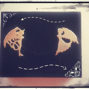 Può includere: Un paio di orecchini dorati, uno a forma di angelo e l'altro a forma di diavolo, su uno sfondo nero.