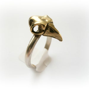 Op de afbeelding: Een zilveren ring met een gouden vogelschedel erop. De schedel is gedetailleerd en heeft een snavel.